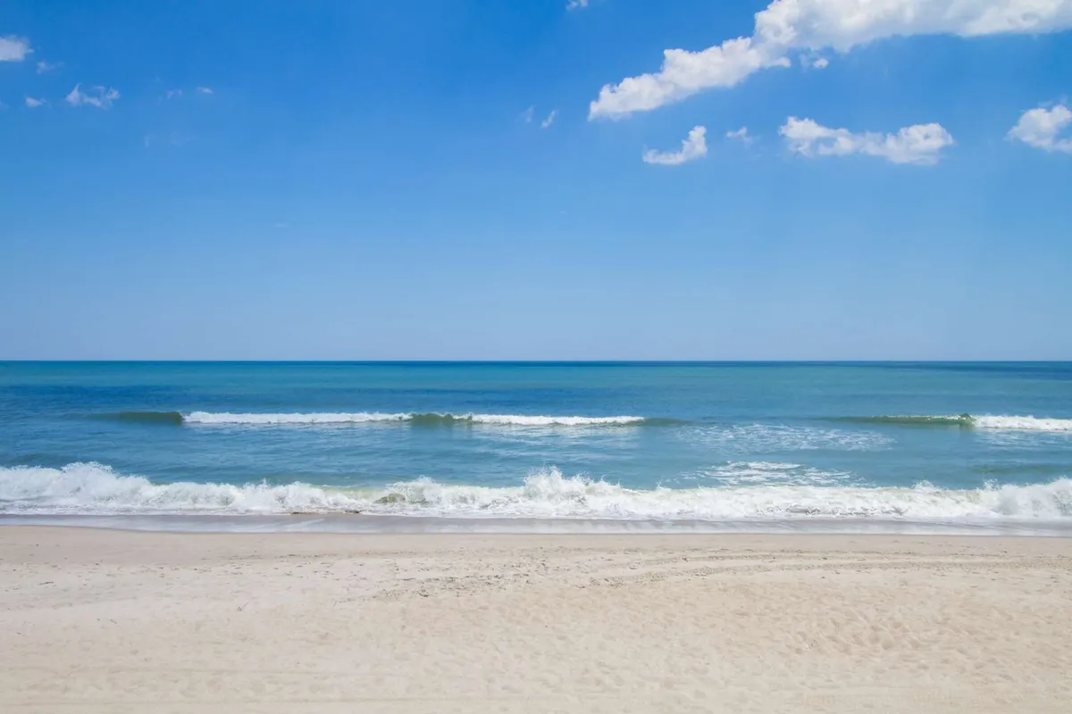 A tranquil beachscape with gentle ocean waves, a wide expanse of sandy beach, and a clear blue sky. The scene is ideal for swimming, sunbathing, and building sandcastles, with no buildings or distractions—just natural beauty and space for family fun.