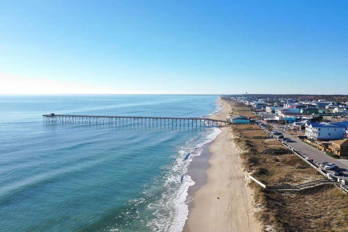 A view of the pier from the beach, with ocean waves and beachfront homes visible. The setting suggests a relaxed walk to the pier, easy access to water sports, and the appealing blend of home comfort and seaside adventure.