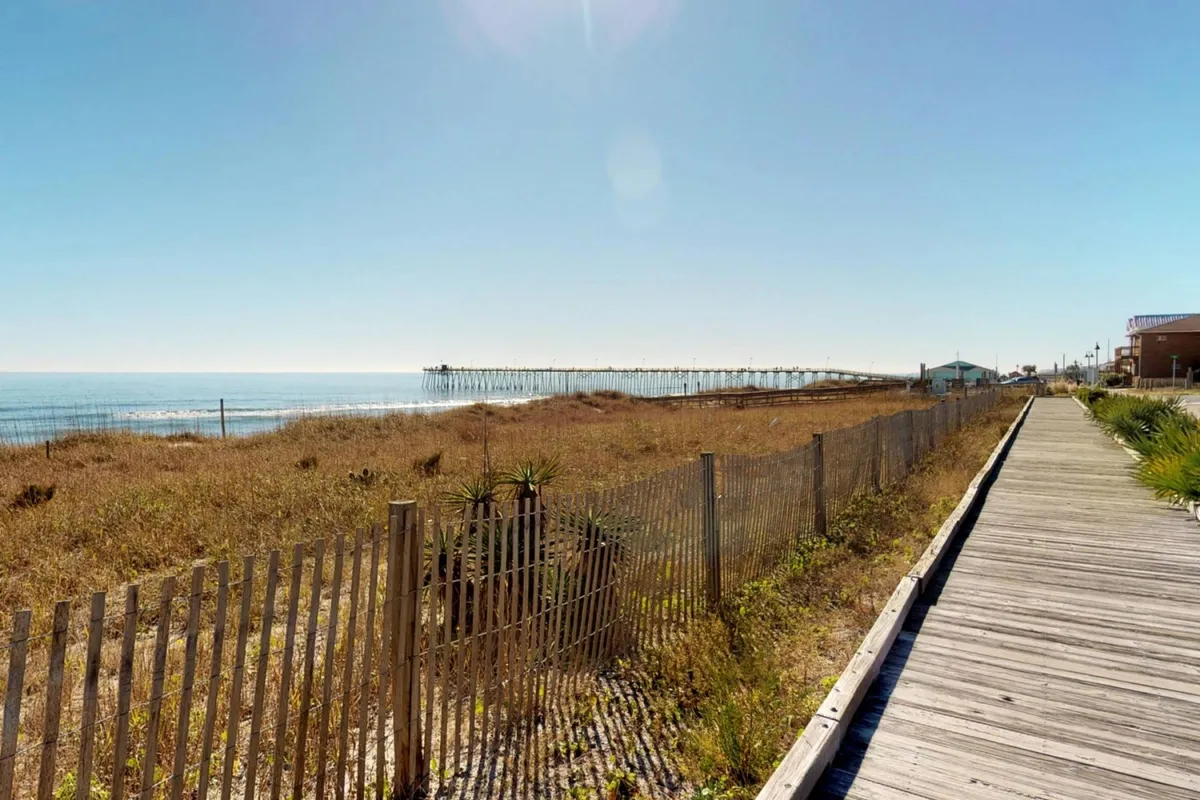 An exterior photograph capturing the long Kure Beach Pier stretching over the ocean, with dune grasses and a boardwalk in the foreground. The view invites thoughts of strolling, fishing, and relaxing by the water, while the gentle waves and early-morning light evoke tranquility and opportunity.