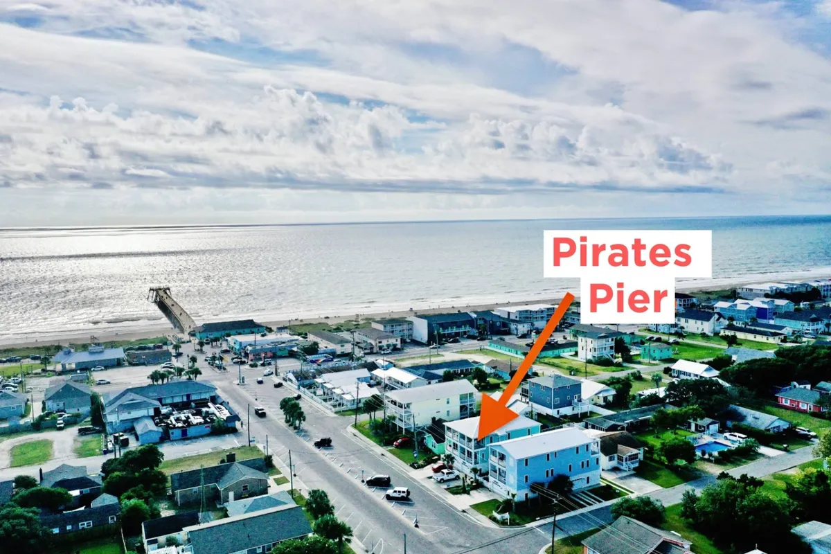 A relaxed exterior view featuring Pirates Pier with the ocean in the background and neighboring residential buildings. The scene captures the essence of the beach area, inviting visitors for outings like fishing and sightseeing at the iconic pier. The setting sun adds a warm glow, indicative of evenings spent by the water.