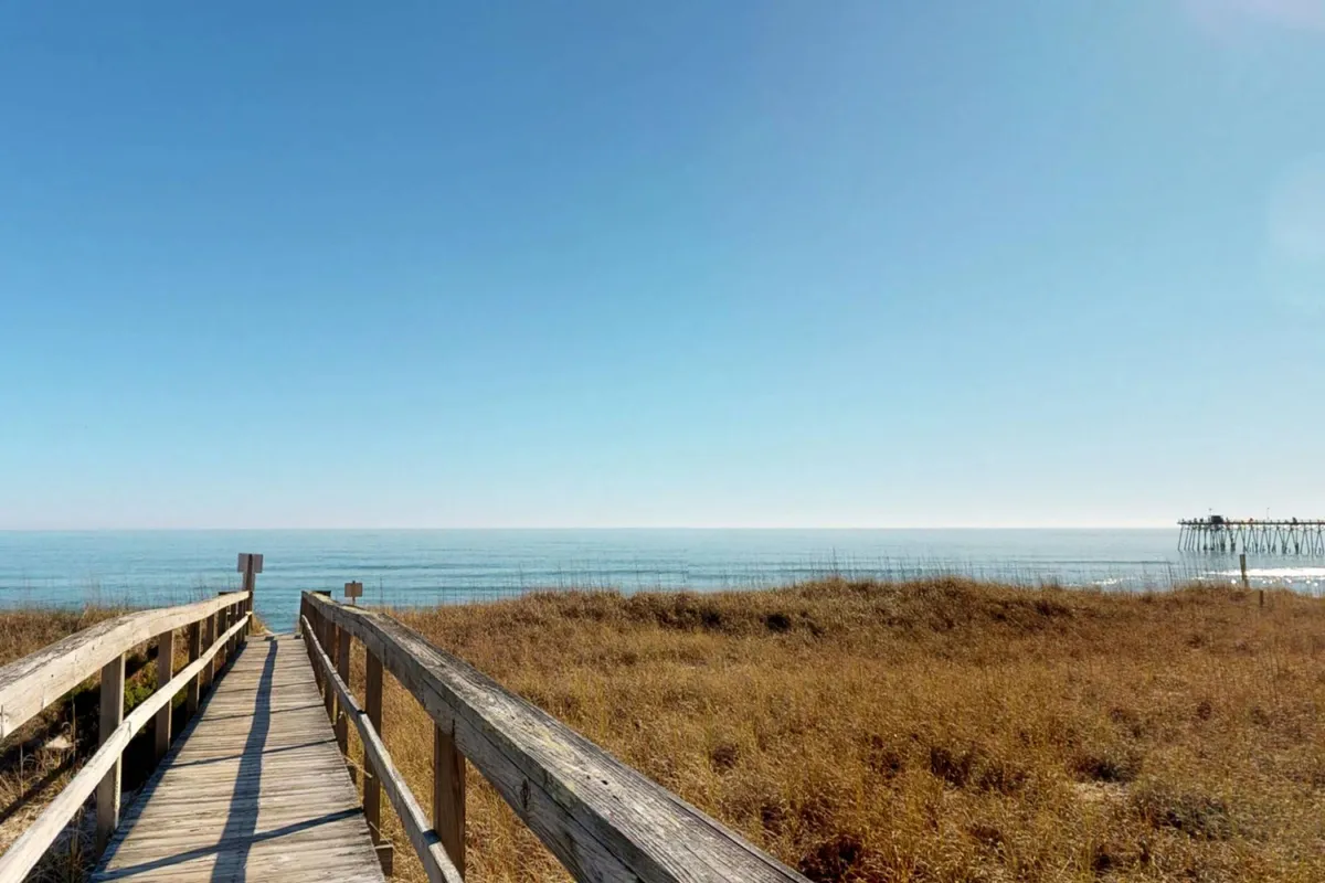 A wooden boardwalk meanders through tall coastal grass, leading toward a panoramic ocean view with a distant pier. The scene is tranquil and inviting, suggesting easy access to the beach and the kind of scenic strolls mentioned in the section.