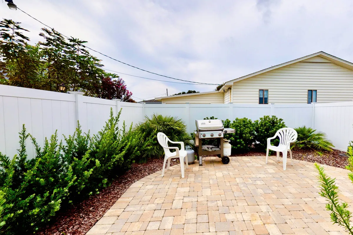 A sunny, paved patio area featuring a modern grill, several white chairs for outdoor seating, lush greenery, and a sturdy fence enclosing the space. The image exudes a sense of privacy and relaxation, inviting guests to enjoy meals and leisure time outside with family or pets.