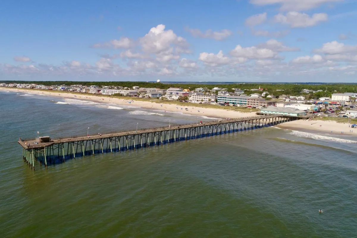 A lively exterior image showing the pier, sandy beach, ocean, and nearby buildings, with people relaxing and exploring the area. The clouds and sunlight create a relaxed, social seaside atmosphere, reinforcing the idea of group outings and fun close to home.