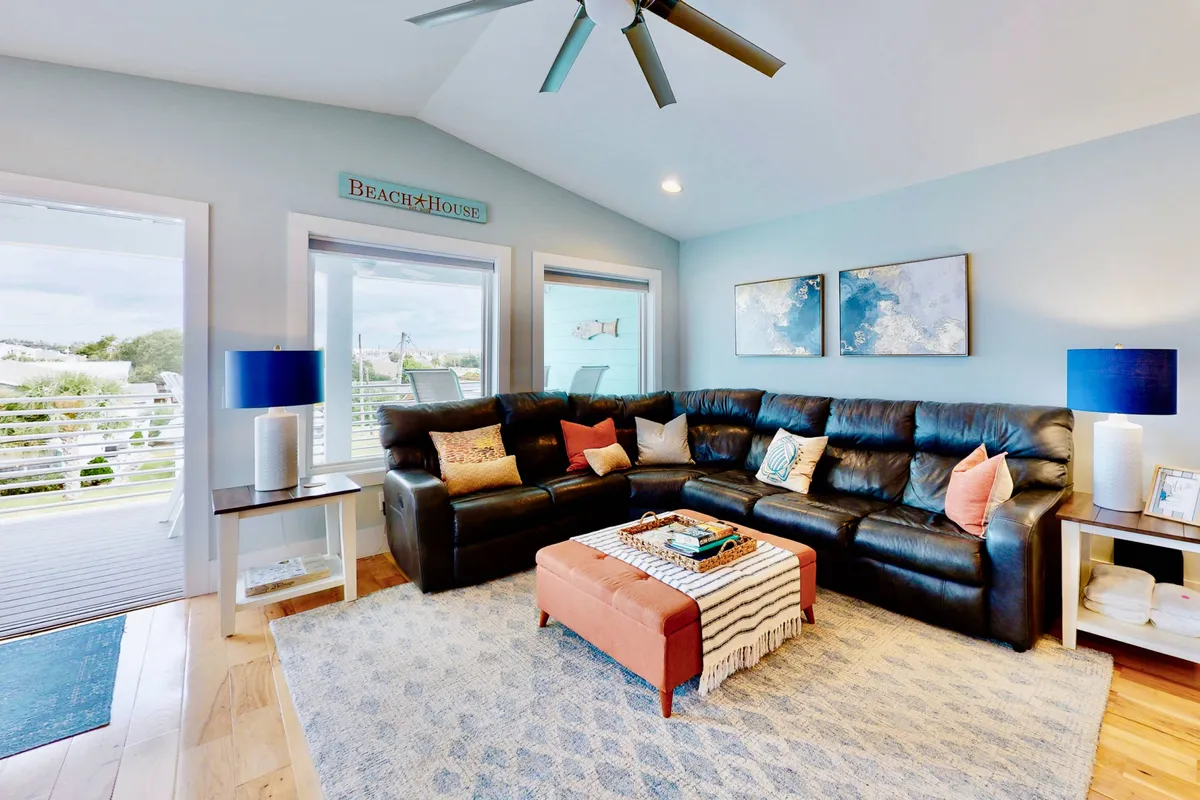 A spacious, open-concept living room featuring a large sectional sofa with colorful throw pillows, a coffee table with stylish decor, and artwork on the walls. Sunlight pours in through expansive windows, offering a glimpse outside and filling the room with warmth and brightness. The comfortable setup encourages gatherings and relaxation.