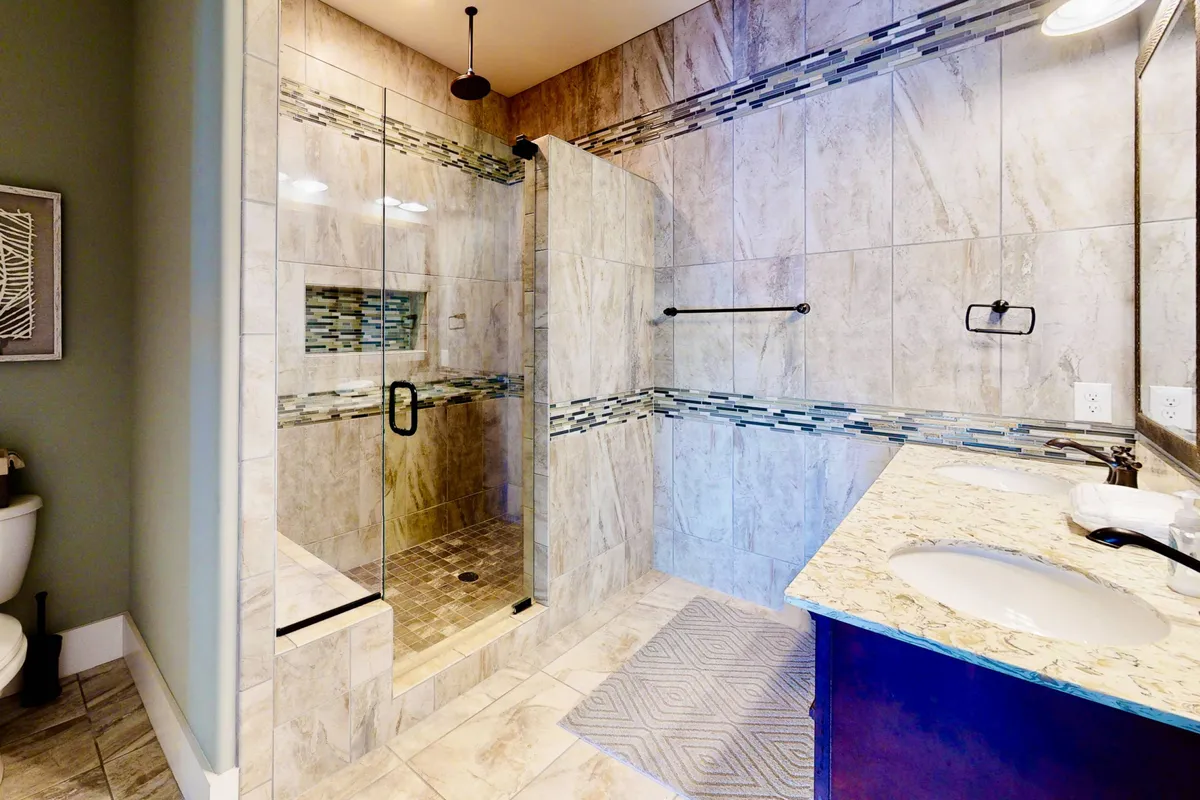 A spacious, modern bathroom with a glass shower enclosure, granite countertop, double sink vanity, decorative tile accents, and a plush rug. The space exudes a spa-like, refreshing ambiance ideal for relaxation and self-care.