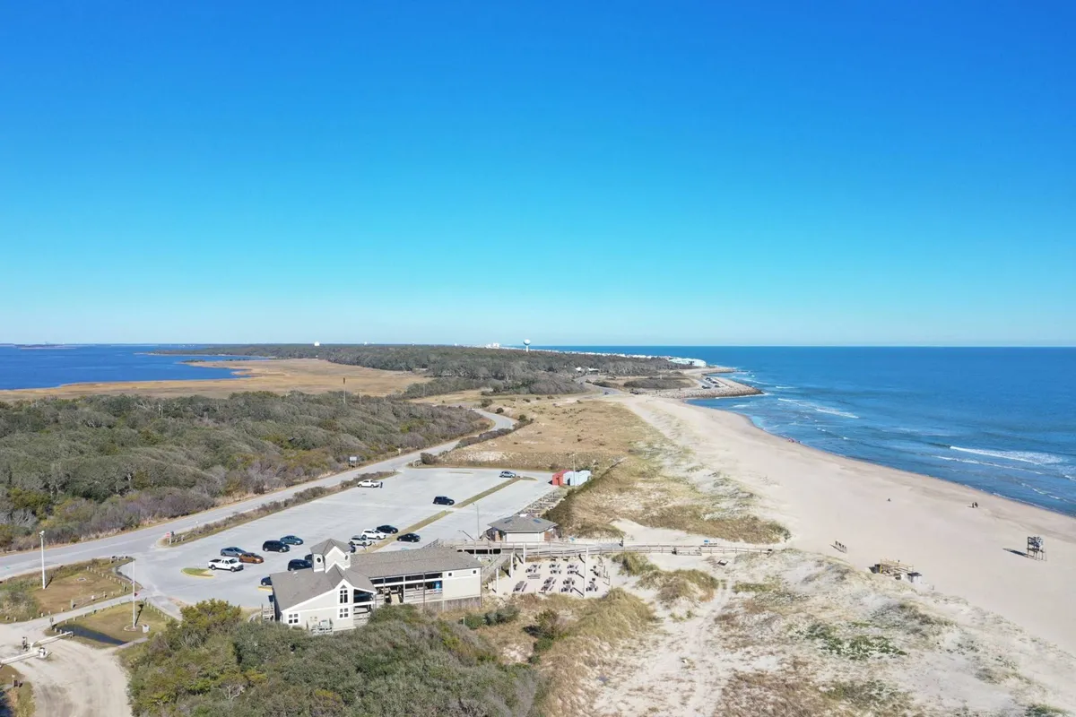 A sweeping view of a sandy beach, calm ocean waves, a nearby restaurant, and natural coastal vegetation. The image captures the relaxed yet upscale vibe of the surroundings, with easy access to fine dining and outdoor leisure.