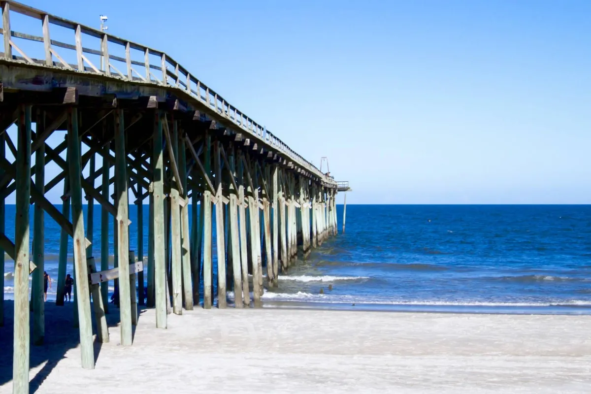 A wide shot of the iconic Kure Beach pier stretching out over the calm ocean, with sandy shoreline and gentle waves in the foreground. The scene is tranquil, inviting visitors to stroll along the pier or watch the sunset. Nearby, the atmosphere hints at the proximity of restaurants and local attractions.