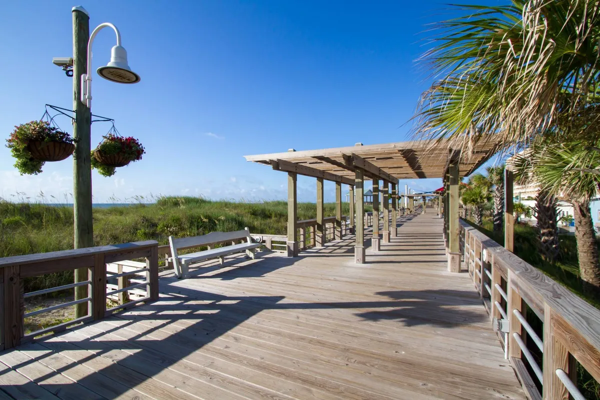 A vibrant exterior shot of a wooden boardwalk adorned with hanging flower baskets, palm trees, and a pergola structure, surrounded by lush greenery. The peaceful setting suggests a pet-friendly neighborhood ideal for leisurely walks and outdoor relaxation.
