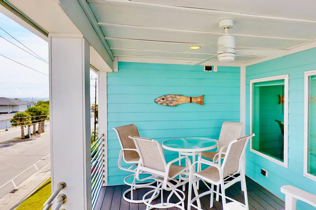 The image showcases the condo's inviting outdoor patio, featuring a cheerful light blue wall, a stylish round table surrounded by bar stools, and playful decorative fish accents. The setup hints at a space perfect for enjoying coffee at sunrise, relaxing in the afternoon sea breeze, or gathering for evening meals under the stars.