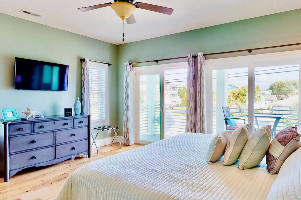 A cozy bedroom with a stylish bed adorned with decorative pillows, a flat-screen TV, a dressing chest, and large windows with curtains. The room is softly lit and feels welcoming for both relaxation and family moments.
