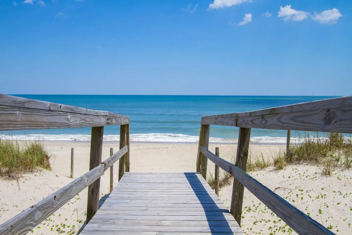A serene exterior shot of a wooden walkway leading directly to the ocean, bordered by sand and sky. The image evokes feelings of morning beach walks, sunset views, and the seamless transition from home to shore.