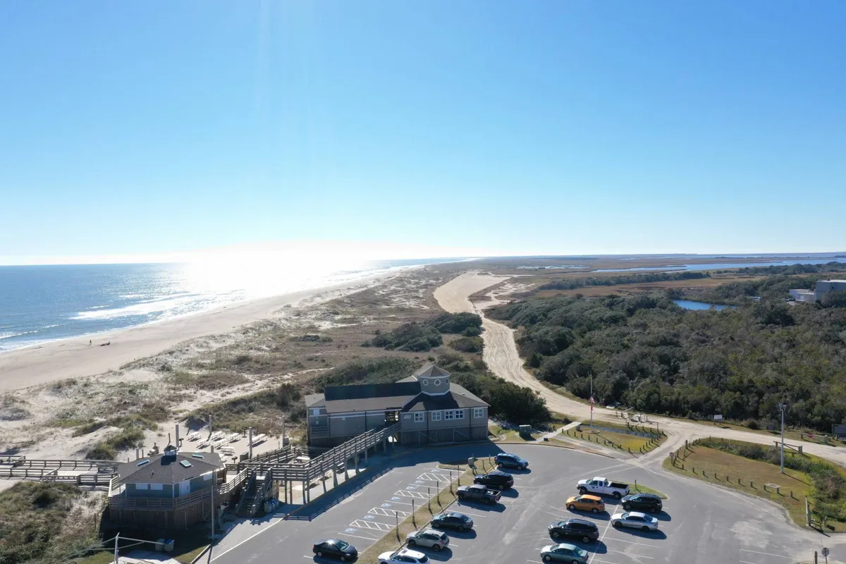 An exterior shot showing the building, adjacent beach, ocean, and a visible parking lot. This scene connects the convenience of plentiful parking with immediate access to the beach, emphasizing the seamless transition from car to sand.