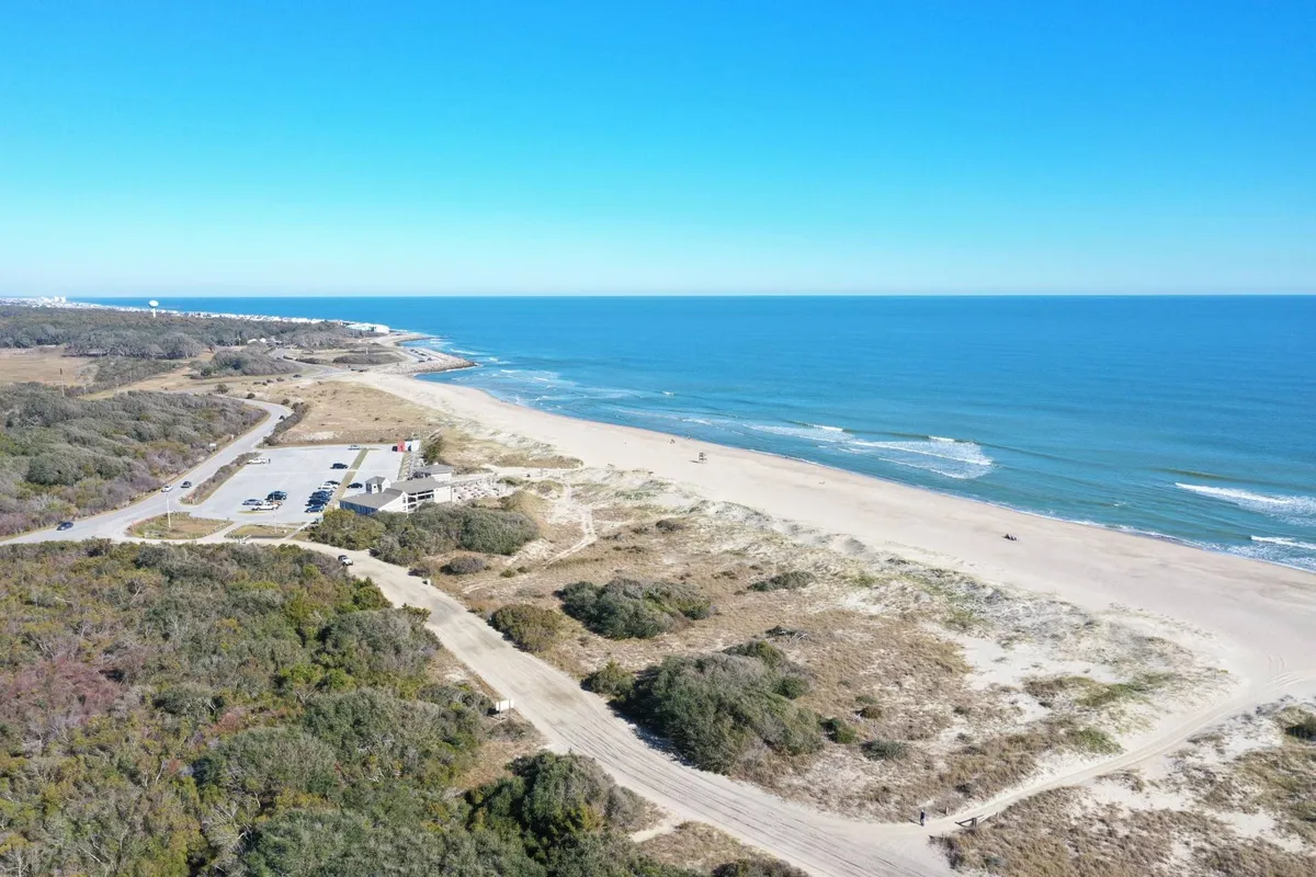 A sweeping view of the beach and ocean framed by coastal vegetation, with a visible parking area that hints at easy access for adventure seekers. The expansive shoreline invites activities like kayaking, paddleboarding, and sunbathing, aligning with the narrative of outdoor exploration.