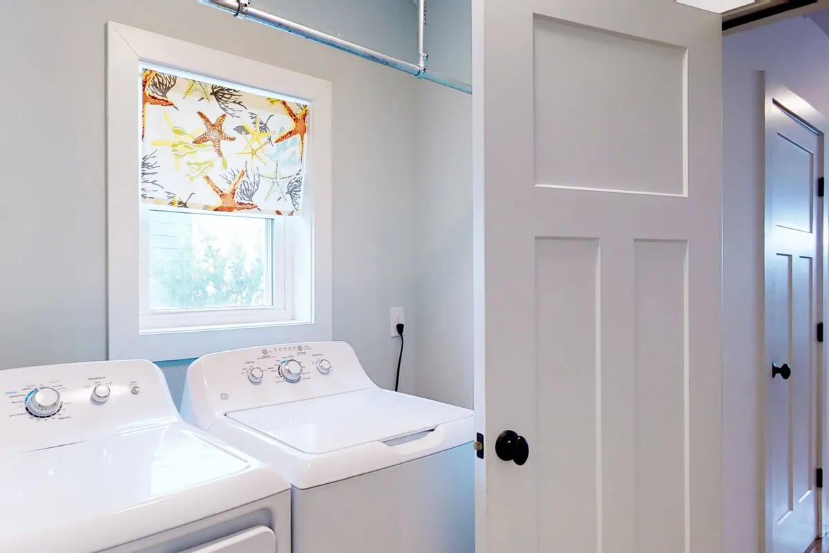 A bright laundry area featuring a modern washing machine and a window adorned with a patterned curtain, allowing natural light to brighten the space. Nearby, shelves or storage space may be used for organizing laundry supplies, creating an efficient and practical environment.